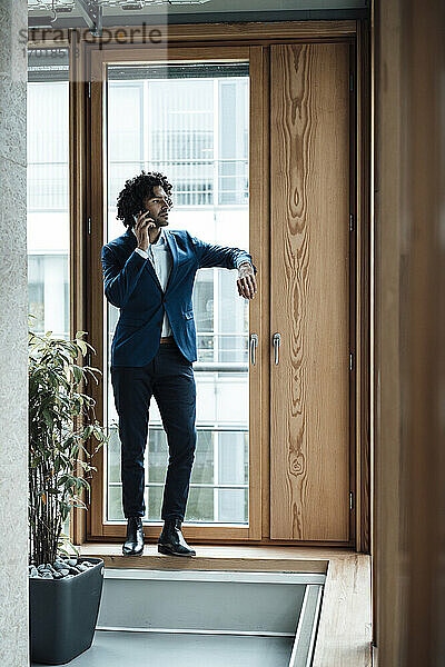 Männlicher Unternehmer  der mit seinem Handy telefoniert  während er im Büro am Fenster steht