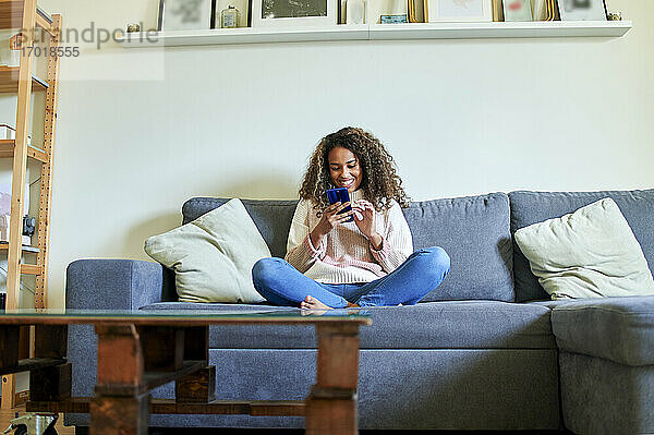 Junge Afro-Frau lächelt  während sie auf dem Sofa im Wohnzimmer ein Mobiltelefon benutzt