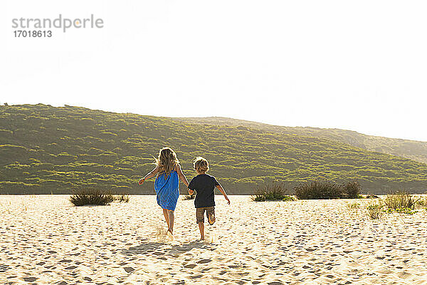 Bruder und Schwester halten sich an den Händen  während sie auf Sand laufen