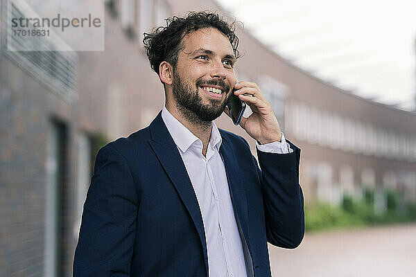 Lächelnder männlicher Unternehmer  der mit seinem Smartphone vor einem Gebäude spricht