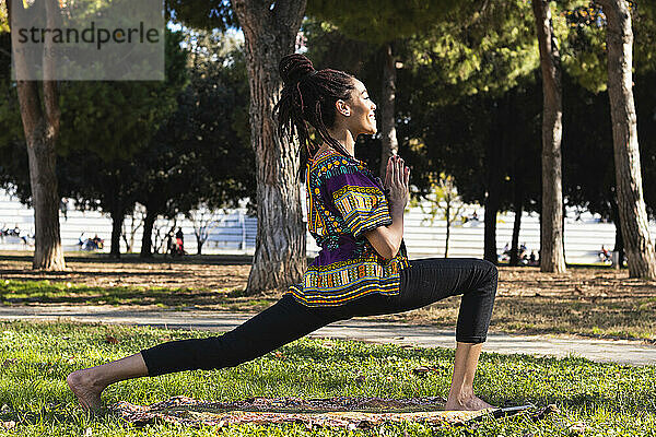 Lächelnde junge Frau übt Yoga im Park an einem sonnigen Tag