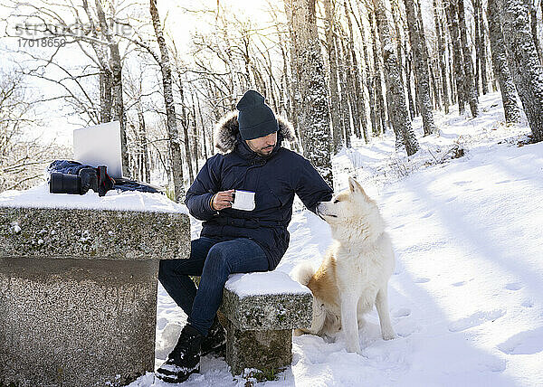 Mittlerer erwachsener Mann mit Kaffeetasse  der seinen Hund streichelt  während er auf einer Felsbank im Schnee sitzt