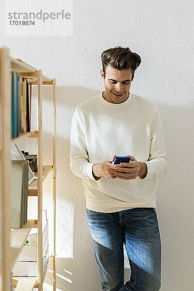 Lächelnder junger Mann  der sein Smartphone an einer weißen Wand neben einem Bücherregal benutzt