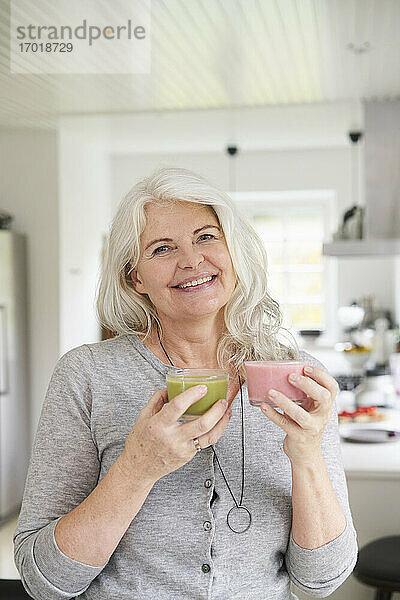 Lächelnde Frau im Ruhestand mit frischen Smoothies in der Hand  während sie in der Küche zu Hause steht