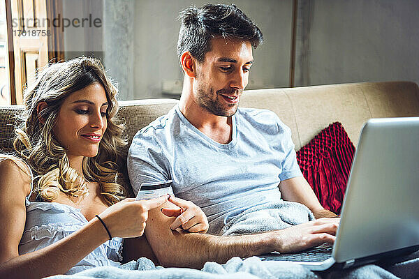 Junges Paar sitzt zu Hause auf der Couch mit Kreditkarte und Laptop