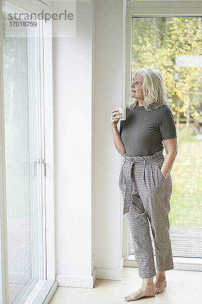 Nachdenkliche Frau im Ruhestand hält eine Tasse in der Hand und schaut durch das Fenster ihres Hauses