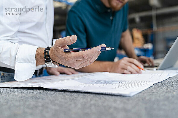 Hand eines Mannes  der einen Stift hält  während er mit einem Mitarbeiter über Dokumente spricht