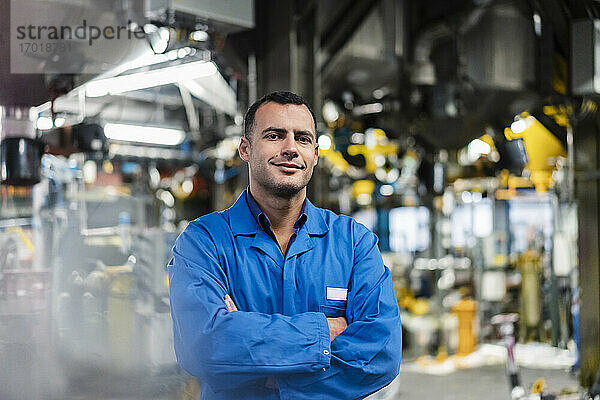 Lächelnder männlicher Ingenieur mit verschränkten Armen in einer Fabrik stehend