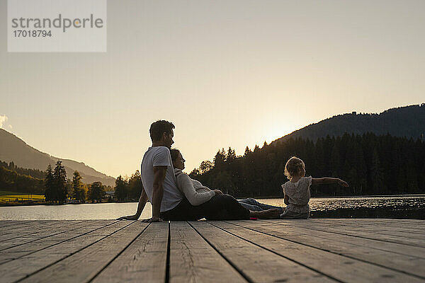 Familie mit kleiner Tochter sitzt bei Sonnenuntergang am Ende eines Stegs am Seeufer zusammen