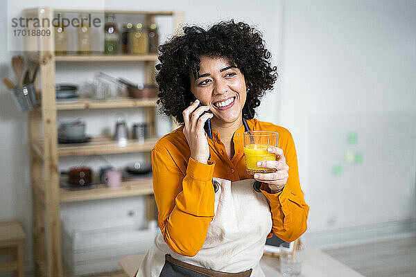 Lächelnde Frau mit Orangensaft im Gespräch auf dem Handy  während sie zu Hause steht
