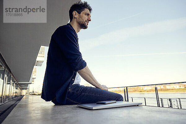 Geschäftsmann mit Blick auf die Aussicht  während er mit Laptop und Mobiltelefon auf einer Stützmauer sitzt