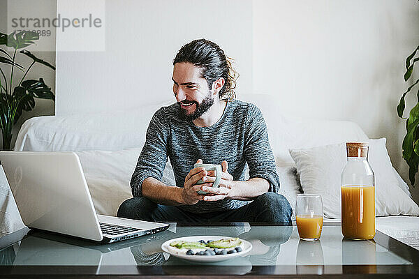 Junger Mann mit Kaffeetasse und Laptop  während er zu Hause auf dem Sofa sitzt und frühstückt