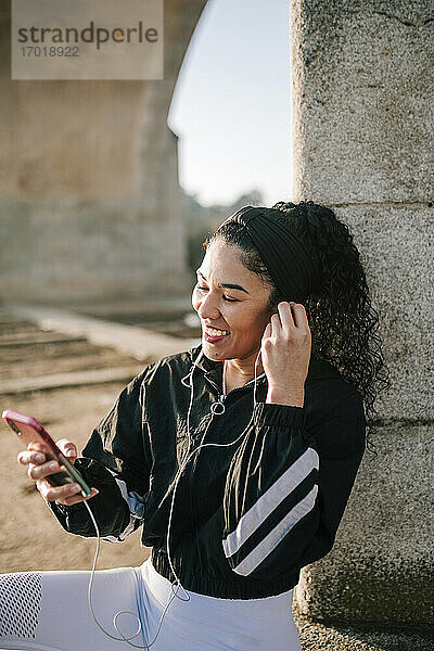 Glückliche Sportlerin  die an einem sonnigen Tag In-Ear-Kopfhörer einstellt  während sie ein Mobiltelefon benutzt