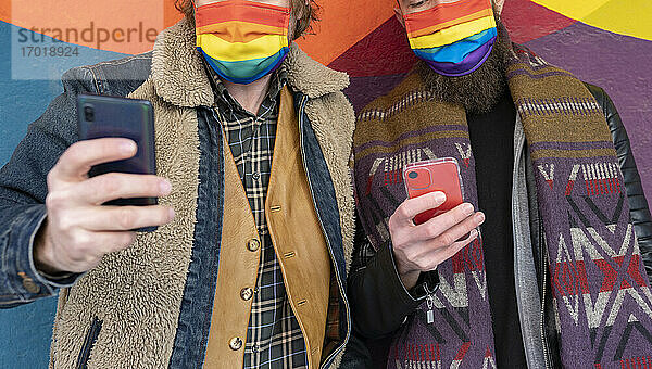 Schwule Freunde mit Regenbogenmasken benutzen Smartphones  während sie an der Wand stehen