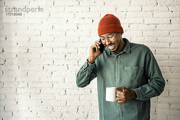 Lächelnder Mann mit Strickmütze telefoniert und hält eine Kaffeetasse an eine weiße Backsteinwand