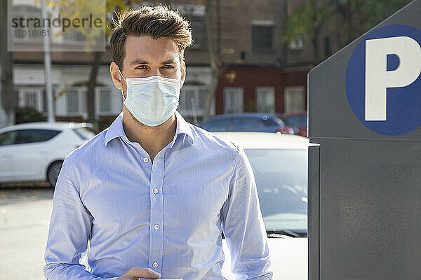 Geschäftsmann mit Gesichtsmaske schaut weg  während er an einer Parkuhr in der Stadt steht