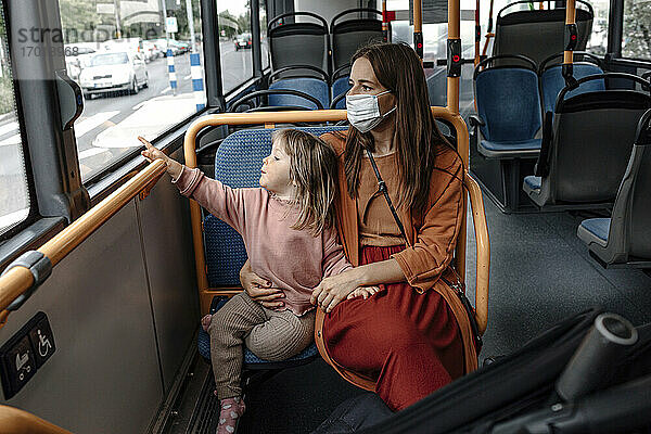 Mutter mit niedlicher Tochter  die auf das Fenster zeigt  während sie mit dem Bus während der COVID-19 reist