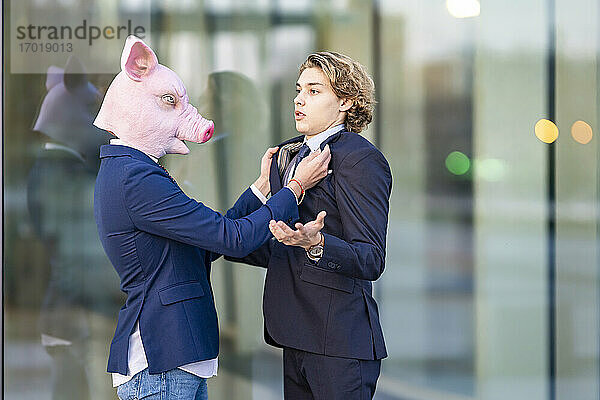 Geschäftsmann mit Schweinemaske hält den Kragen eines männlichen Kollegen an einer Glaswand