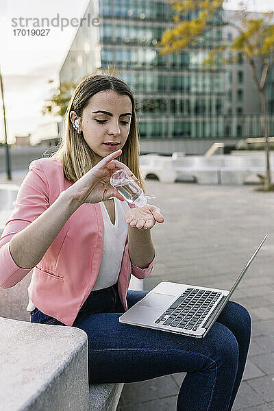 Spanien  Barcelona  Junge Geschäftsfrau mit Laptop benutzt Handdesinfektionsmittel im Freien