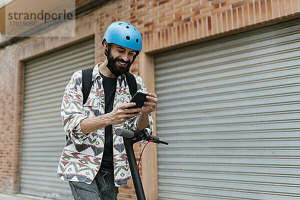 Junger Mann mit elektrischem Tretroller  der sein Smartphone benutzt  während er vor einem Fensterladen steht