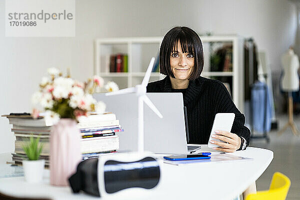 Junge Frau mit Laptop und Handy in der Hand  während sie im Studierzimmer lernt