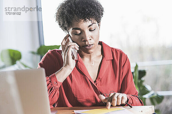 Frau  die am Schreibtisch mit einem Handy telefoniert und auf Dokumente schaut