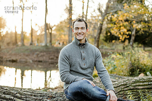 Lächelnder erwachsener Mann sitzt auf einem Baumstamm in einem öffentlichen Park