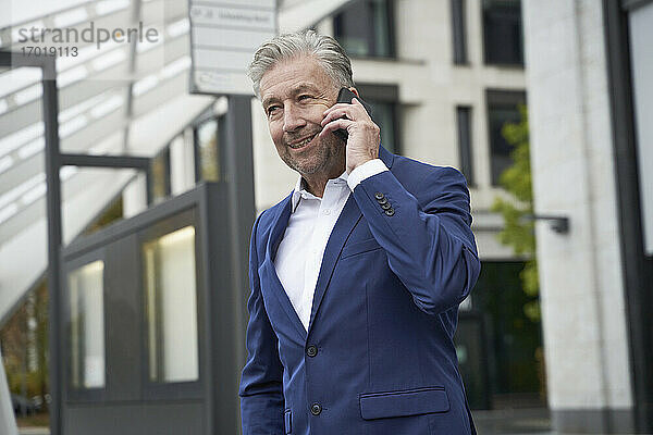 Lächelnder Geschäftsmann  der mit seinem Handy telefoniert  während er an einer Bushaltestelle in der Stadt wartet