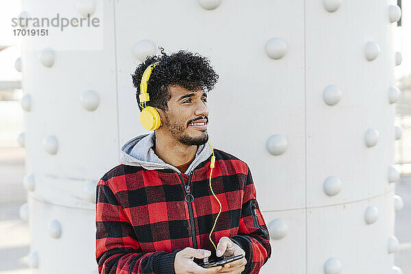 Junger Mann mit Kopfhörern  der ein Mobiltelefon benutzt  während er an der Wand steht