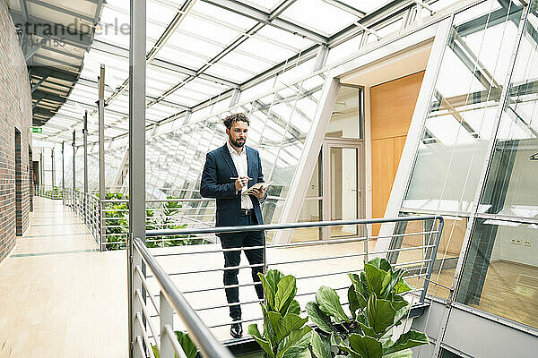 Männlicher Unternehmer mit digitalem Tablet  der eine Pflanze im Flur eines Büros betrachtet