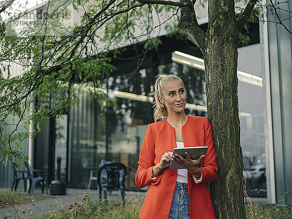 Geschäftsfrau mit digitalem Tablet  die wegschaut  während sie an einem Baum vor einem Bürogebäude steht