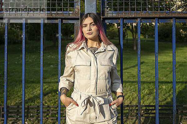 Stilvolle junge Frau mit Händen in den Taschen am Zaun an einem sonnigen Tag