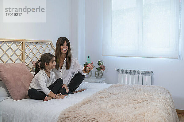 Lächelnde Mutter und Tochter bei einem Videogespräch mit dem Vater über ein Smartphone im Schlafzimmer