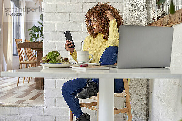 Lächelnde weibliche Fachkraft mit Hand im Haar  die in einem Café ein Mobiltelefon benutzt