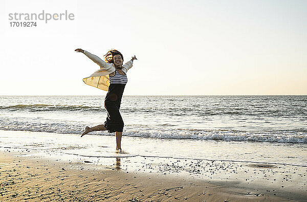 Unbeschwerte junge Frau läuft am Strand gegen den klaren Himmel bei Sonnenuntergang