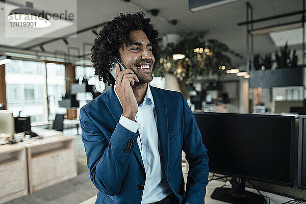 Lächelnder männlicher Jungunternehmer  der mit seinem Smartphone telefoniert und seinen Blick vom Arbeitsplatz abwendet