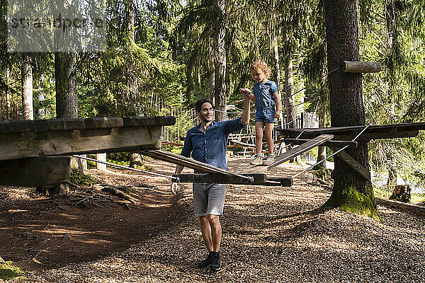 Vater hilft seiner kleinen Tochter beim Gehen über eine kleine Hängebrücke auf einem Hindernisparcours im Wald