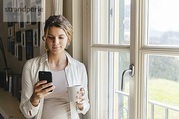 Junge schwangere Frau benutzt ihr Smartphone am Fenster zu Hause