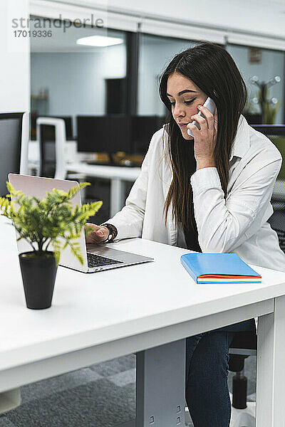 Unternehmerin arbeitet am Laptop  während sie einen Anruf am Schreibtisch im Büro entgegennimmt