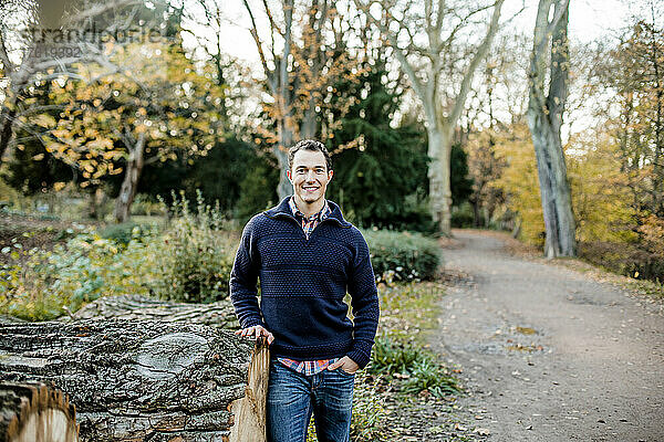 Lächelnder gut aussehender Mann mit Hand in der Tasche vor Bäumen in einem öffentlichen Park stehend