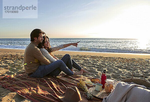 Lächelnde Frau zeigt mit dem Finger auf das Meer  während sie mit einem Mann am Strand sitzt
