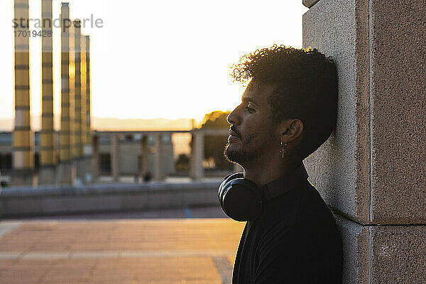 Nachdenklicher junger Mann mit Kopfhörern an der Wand lehnend gegen den klaren Himmel bei Sonnenuntergang