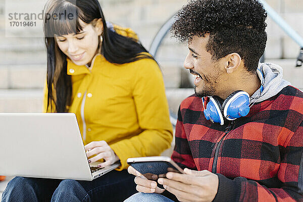 Mann mit digitalem Tablet lächelt  während er neben einem Freund sitzt  der einen Laptop im Freien benutzt