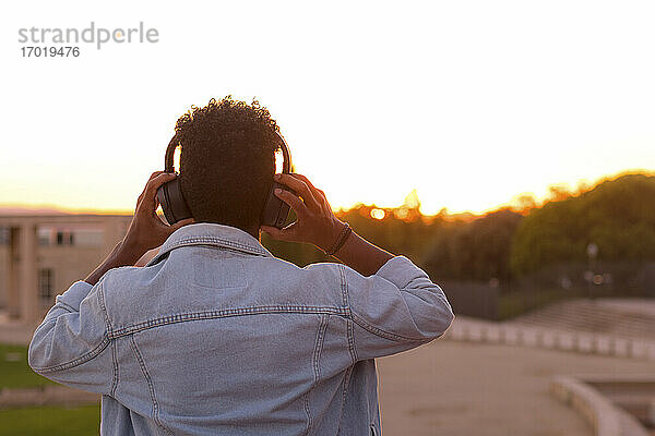 Junger Mann hört Musik über Kopfhörer im Park gegen den klaren Himmel bei Sonnenuntergang
