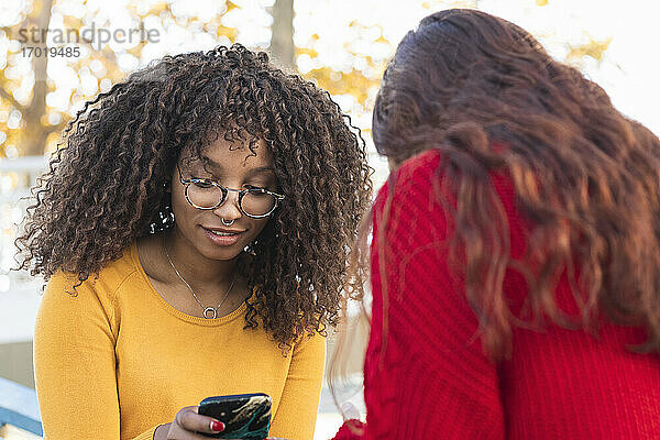 Lächelnde Afro-Frau mit Freundin  die in einem öffentlichen Park ein Mobiltelefon benutzt