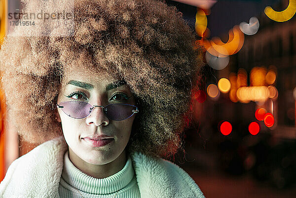 Nahaufnahme einer stilvollen Frau mit Afro-Haar und Sonnenbrille in einer beleuchteten Stadt bei Nacht