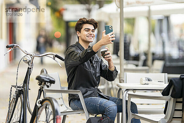 Lächelnder gut aussehender Mann  der ein Selfie mit einer Kaffeetasse durch ein Smartphone macht  während er in einem Straßencafé in der Stadt sitzt