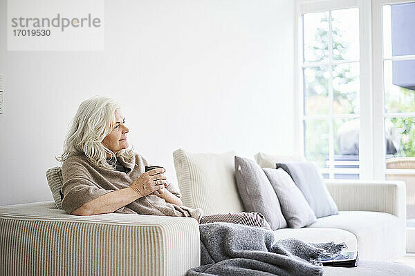 Nachdenkliche Frau mit langen weißen Haaren hält eine Kaffeetasse  während sie auf dem Sofa in einer Wohnung sitzt
