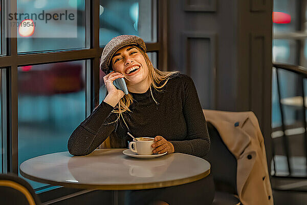 Frau lacht mit Kaffeetasse  während sie in einem Café mit ihrem Smartphone telefoniert