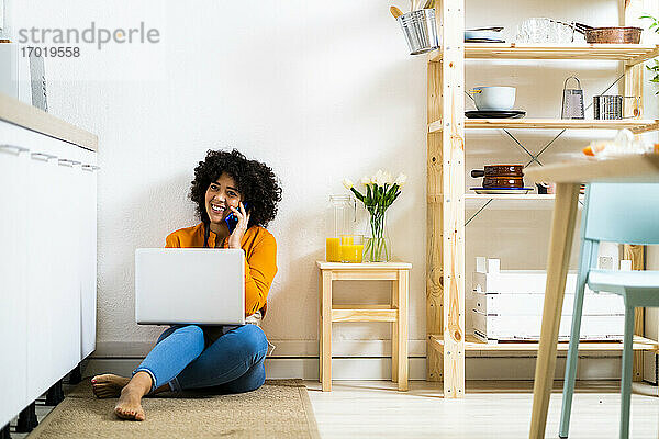 Lächelnde junge Frau mit Laptop  die mit ihrem Handy telefoniert  während sie zu Hause auf dem Boden sitzt